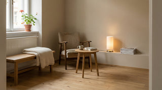 Béžové křeslo s dřevěnými područkami stojící u kulatého dřevěného stolku, na kterém je hrnek a kniha.